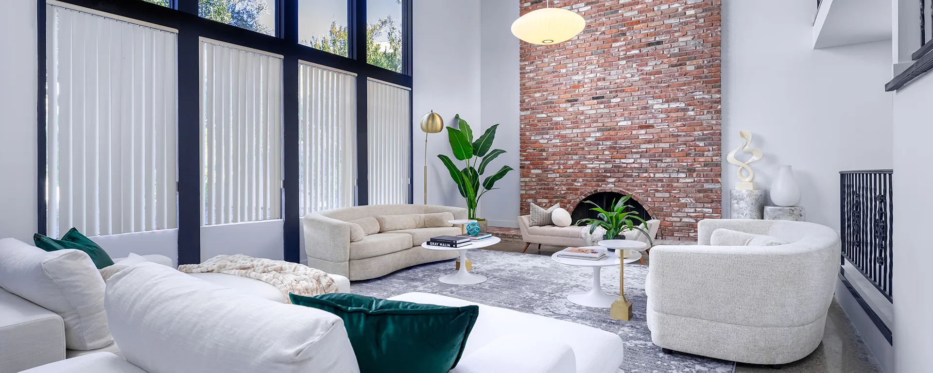 A bright living area with a tall exposed brick wall, curved sofas, and large windows framed in black.