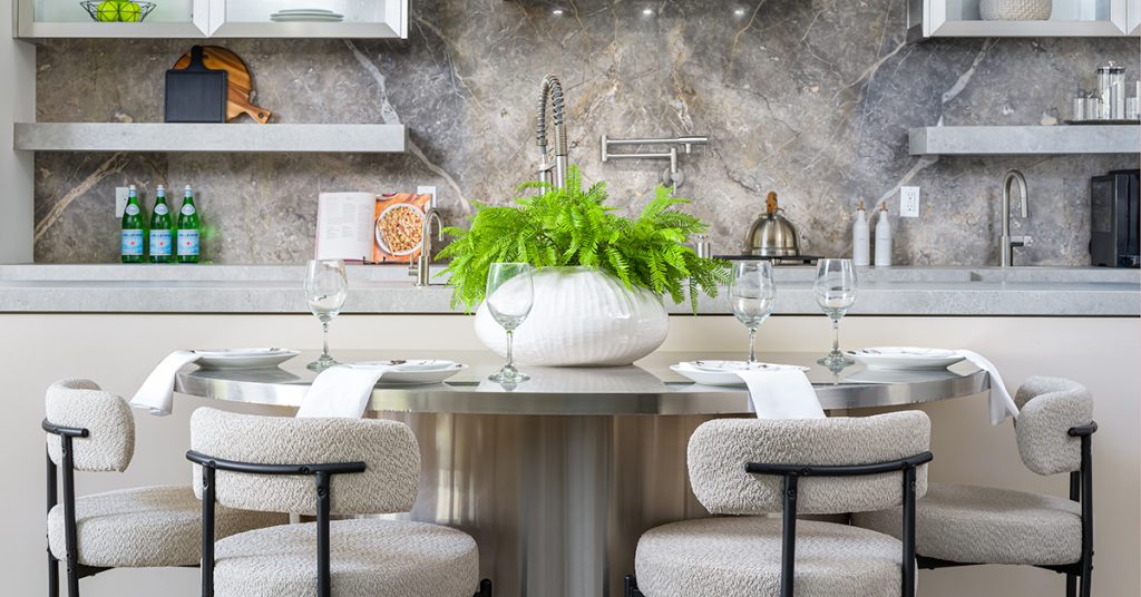 A round metal dining table centered by a white vase of vibrant ferns, surrounded by plush chairs. A gray marble backsplash and open shelving define the kitchen area.