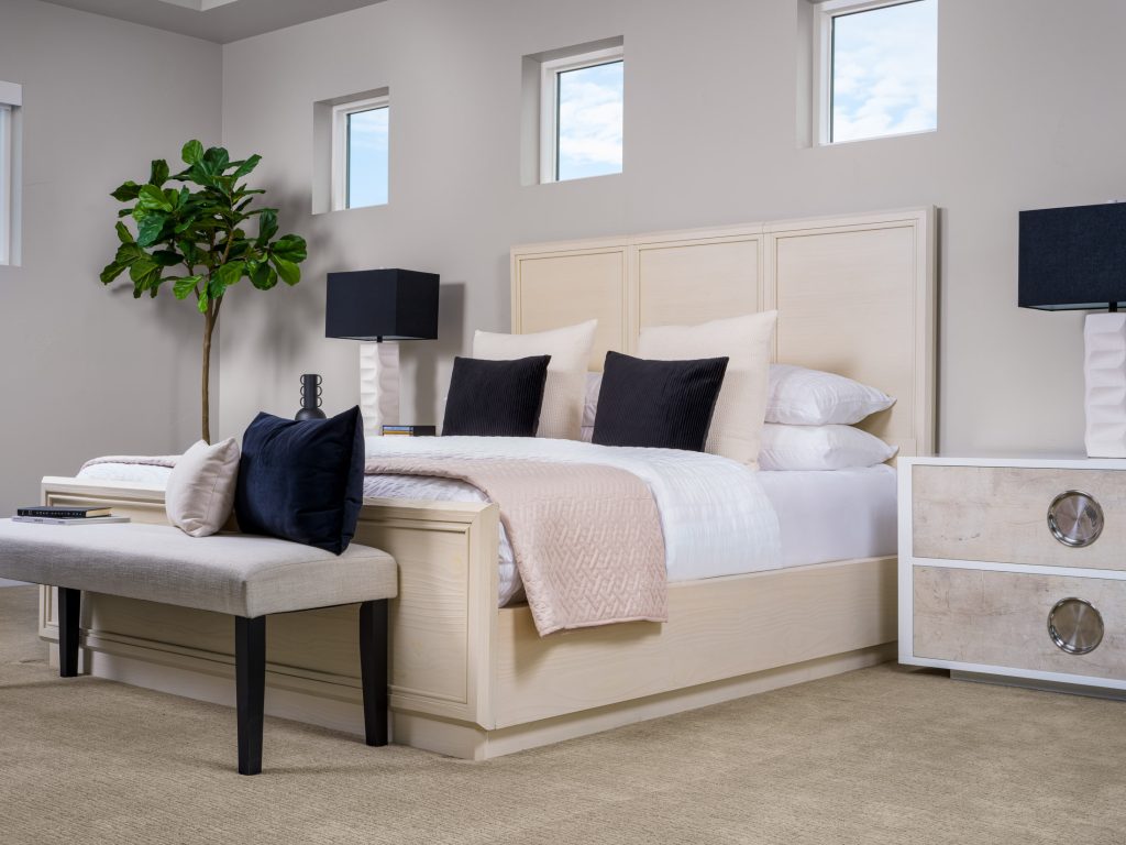 A light wood bed with layered neutral linens and black accent pillows, a gray bench at the foot, and a fiddle leaf fig in the corner.