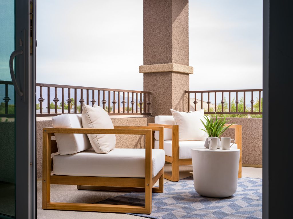 A covered outdoor balcony with wooden chairs featuring white cushions, a small round table, and two mugs beside a green potted plant.