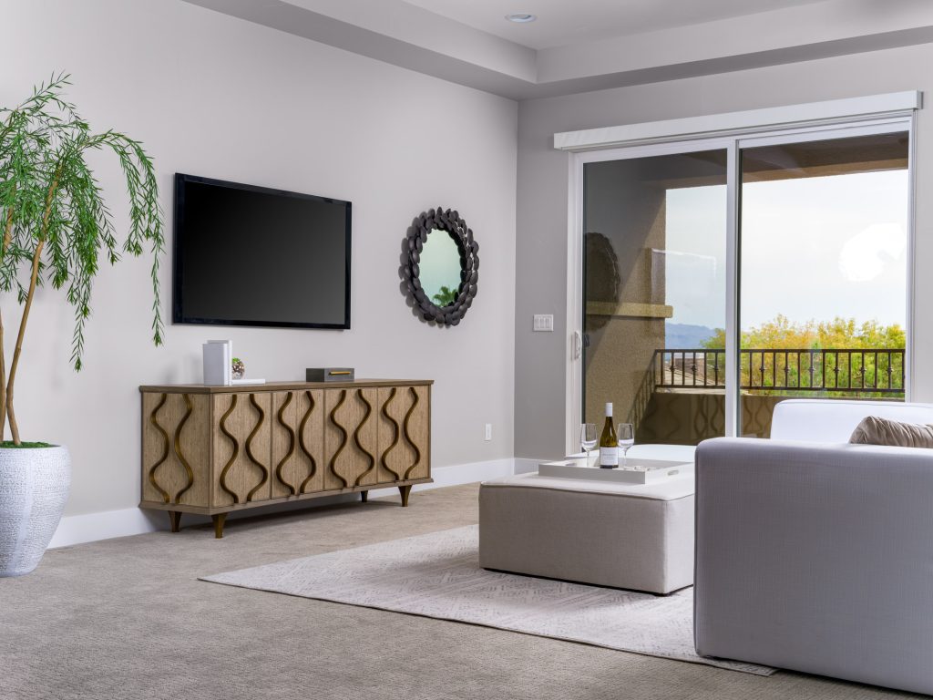 A wooden console with wavy front panels beneath a wall-mounted TV, next to a black circular mirror and a large potted plant, near sliding glass doors.