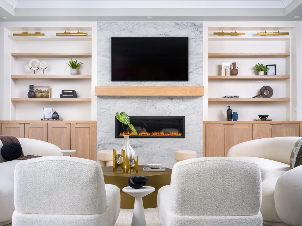 A modern living area with a marble fireplace surrounded by built-in shelves featuring neutral decor. Curved white seating is arranged around a round gold coffee table.