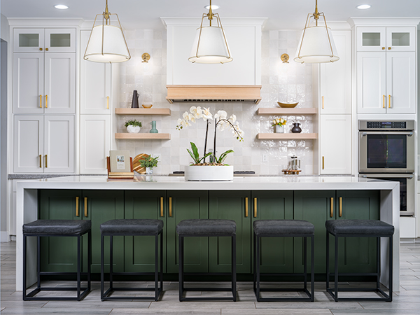 A stunning kitchen featured on Stately’s About Us page, showcasing a large island with a green base and sleek gold hardware, paired with modern black stools. The space is accented by white cabinetry, open wooden shelves, brass light fixtures, and a center orchid arrangement, embodying Stately’s luxury home staging and interior design expertise.