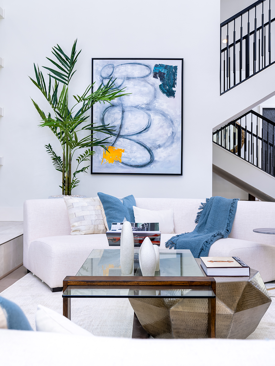 Living room with a white sectional sofa, a glass coffee table, and a large abstract painting on the wall. There is also a tall green plant in the corner, and a staircase with black railings is visible in the background, creating a modern and stylish space.