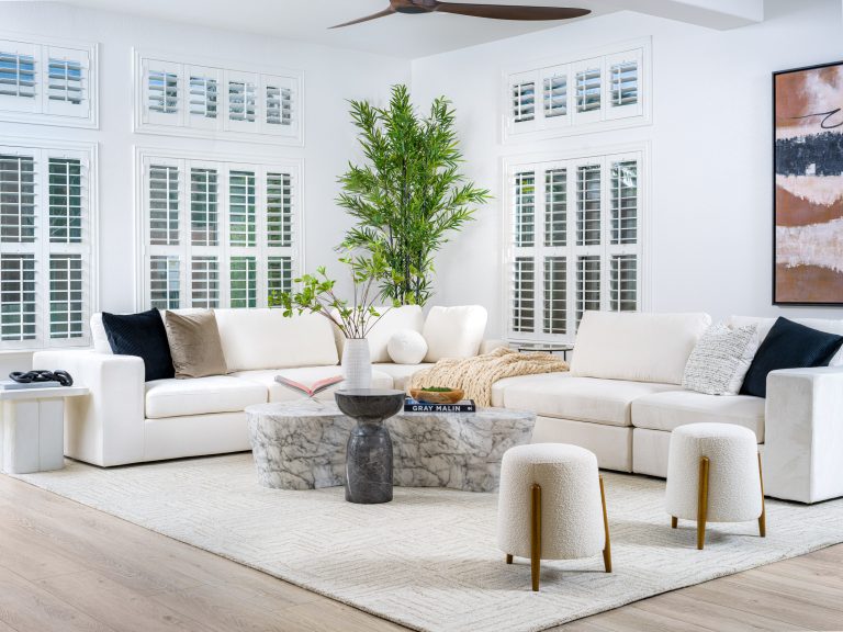 A spacious living room, styled by Contemporary Elegance Home Staging, featuring a white L-shaped sectional, a sculptural marble coffee table, two round stools with wooden legs, and tall shuttered windows.
