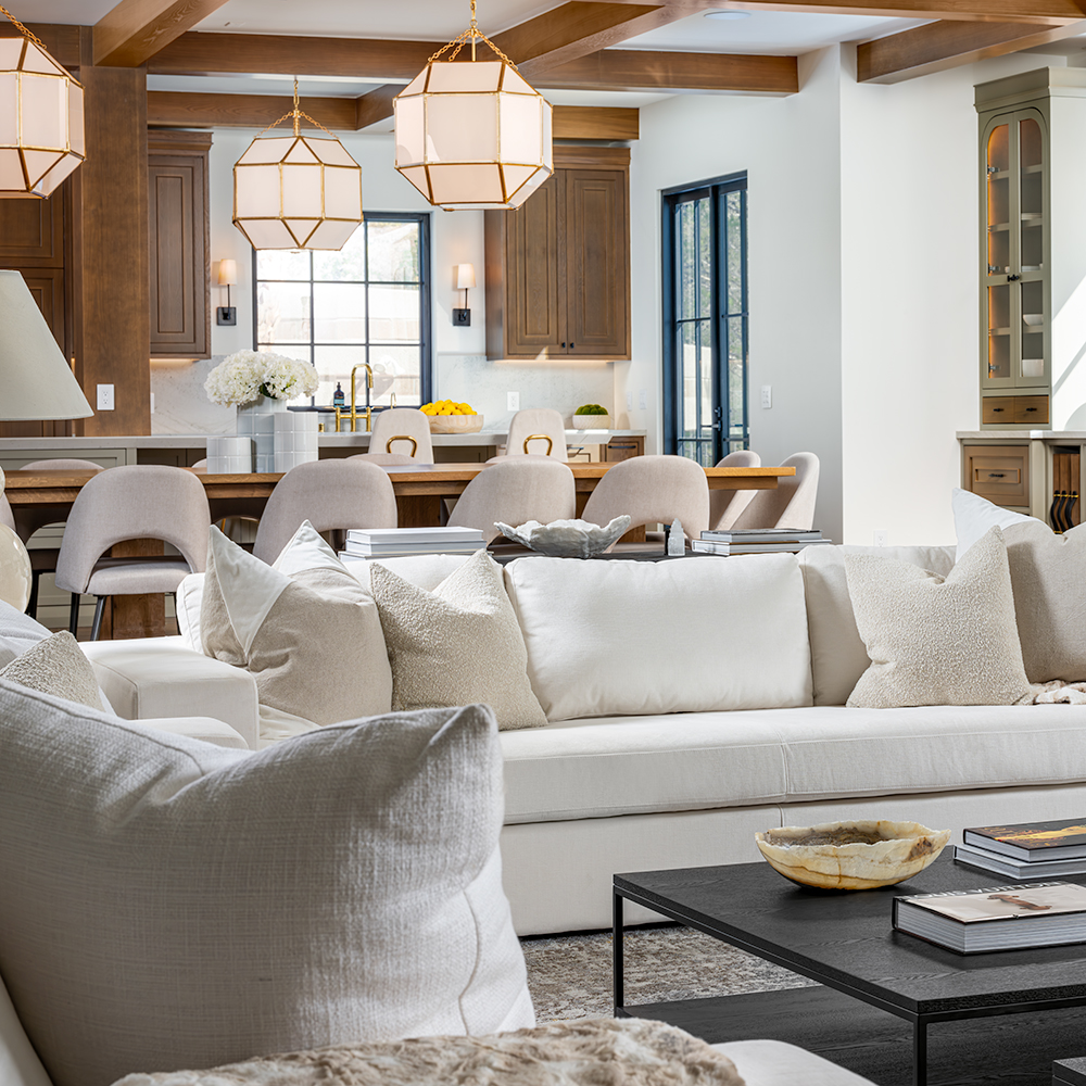 A cozy yet elegant living space featured on Stately’s About Us page, positioned next to testimonials. The design includes a plush white sectional, neutral pillows, a sleek black coffee table, and a backdrop of a sophisticated kitchen with warm wooden cabinetry, gold-accented light fixtures, and modern barstools, reflecting Stately’s dedication to luxurious and welcoming interiors.