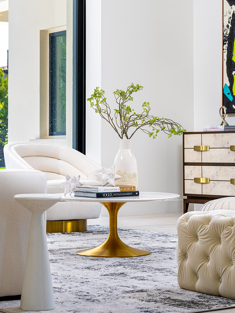 Modern living room with white armchairs, a round white table with a gold base, and a vase with green branches. A large window provides natural light, and a white and gray rug adds texture to the space, creating a bright and inviting atmosphere.