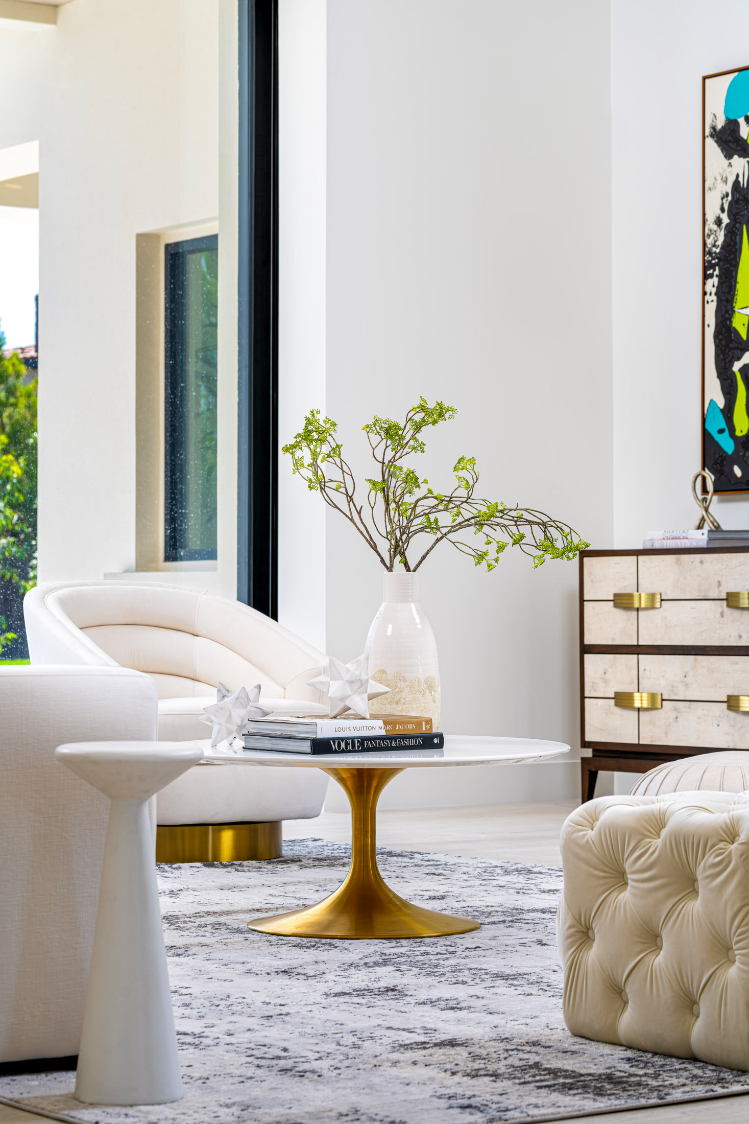 A bright living room with neutral chairs, a gold coffee table, and a decorative branch arrangement in a white vase.