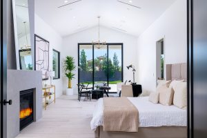 A spacious bedroom with a vaulted ceiling, neutral bedding, and a modern fireplace. Black chairs and a small table sit by large sliding doors leading outside.