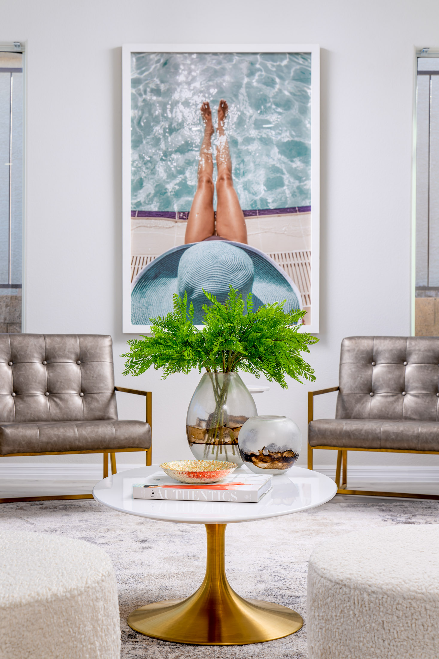 A large photo of legs by a pool hangs above two metallic chairs, with a round gold coffee table and a green plant arrangement in front.