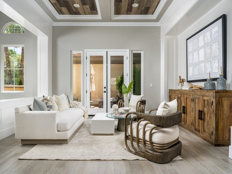 A bright living room, styled by Modern Rustic Home Staging, with wood coffered ceilings, a white sofa, rattan chairs, and glass doors leading outside.