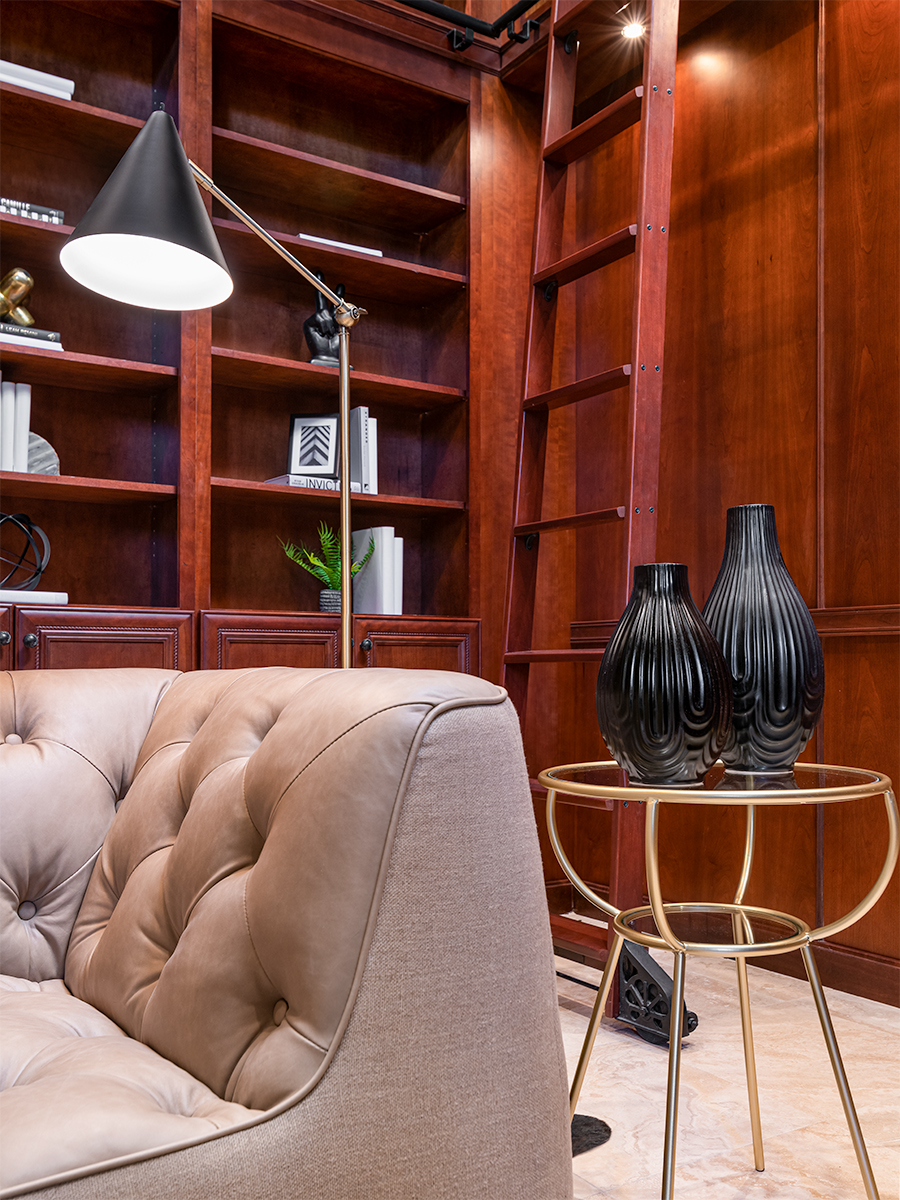 Corner of a library or study with a tufted leather chair, a round side table with decorative vases, and wooden bookshelves filled with books and decorative items. A floor lamp provides light, creating a cozy and inviting space for reading or studying.