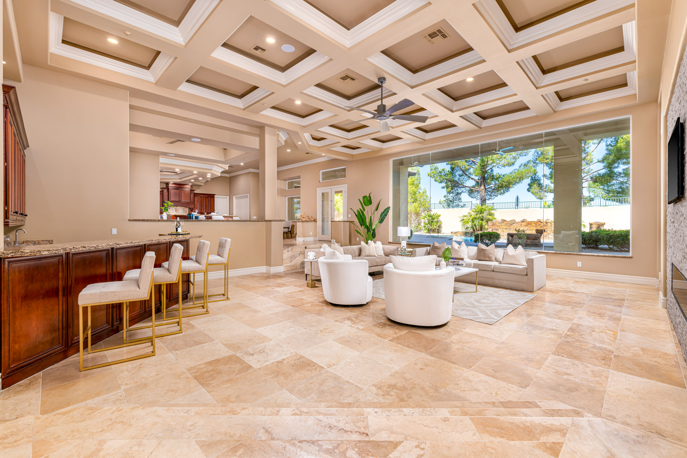A spacious living area with a coffered ceiling, a granite bar, neutral furnishings, and large windows overlooking a pool.
