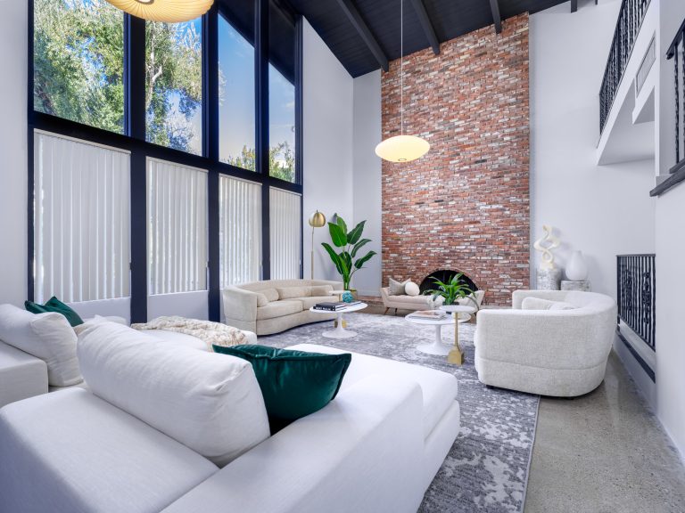 A bright living room featuring a towering brick fireplace, floor-to-ceiling windows, and curved neutral sofas under a high black ceiling.