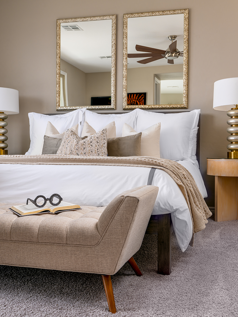 Bedroom with a neatly made bed featuring white linens and beige accent pillows. A beige bench is at the foot of the bed, and two gold-framed mirrors hang above it. A bedside table with a lamp is visible, and the room has a neutral color palette, creating a calming and elegant atmosphere.