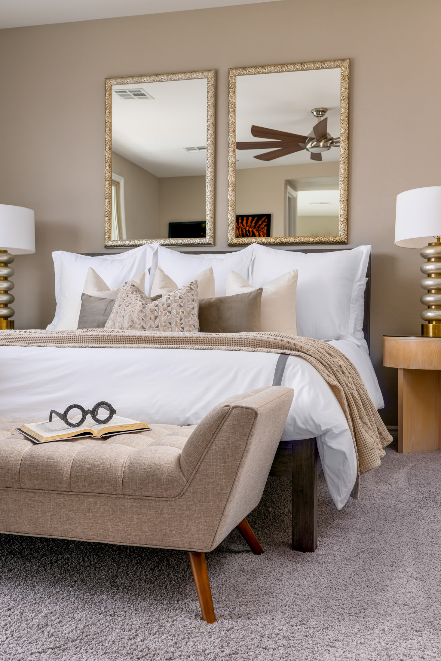 A neutral bedroom with a tufted bench, gold-framed mirrors, white bedding, and layered pillows.