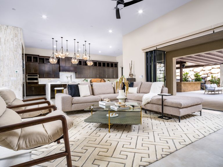 A light-toned sectional sofa, expertly styled by Sophisticated Home Staging, faces a modern kitchen with dark cabinetry and geometric pendant lights. Large sliding doors open to a covered patio.