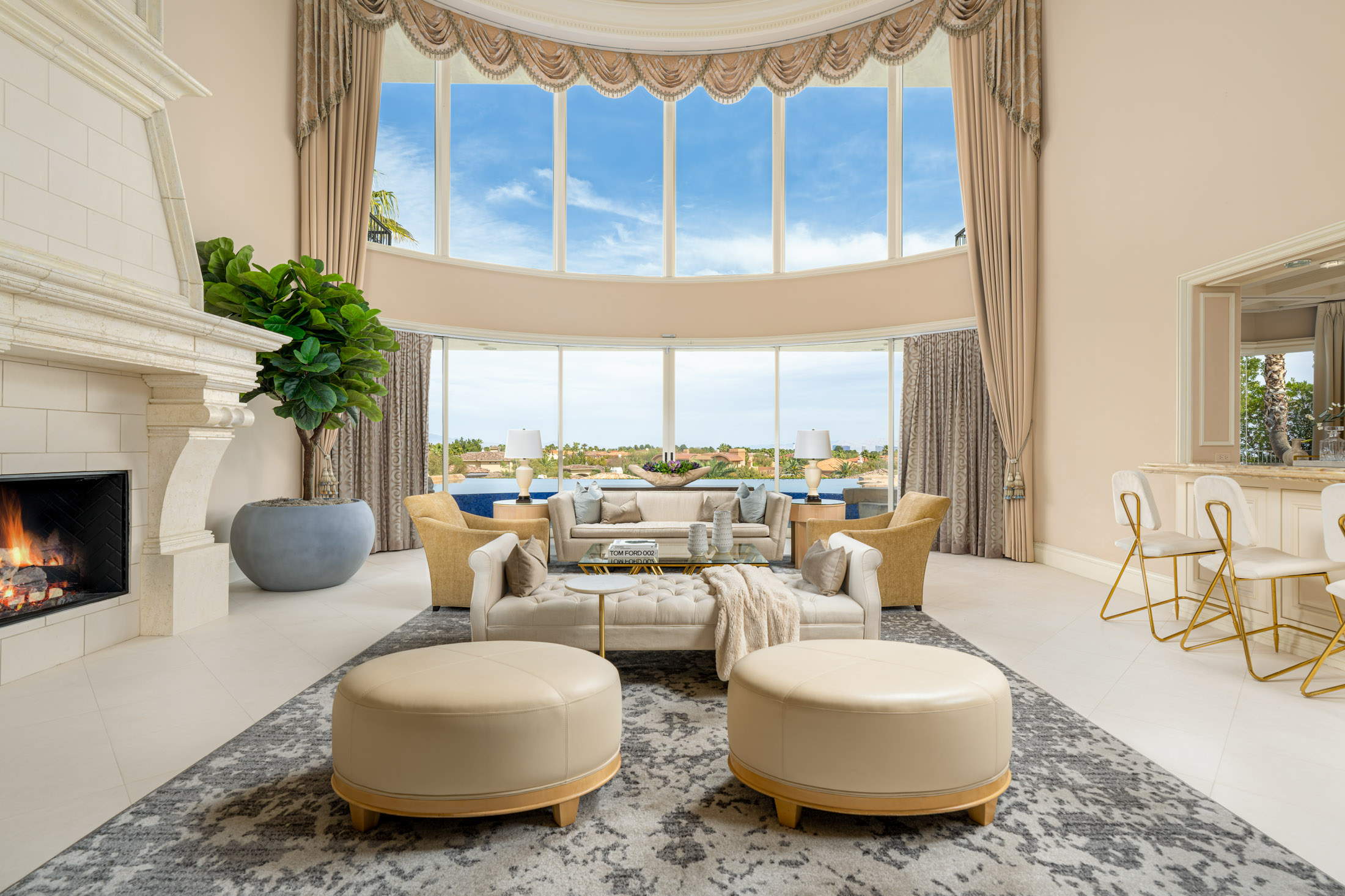 A spacious living room with a curved wall of tall windows, a grand stone fireplace on one side, and plush neutral seating. Large potted greenery and round ottomans complete the open layout.