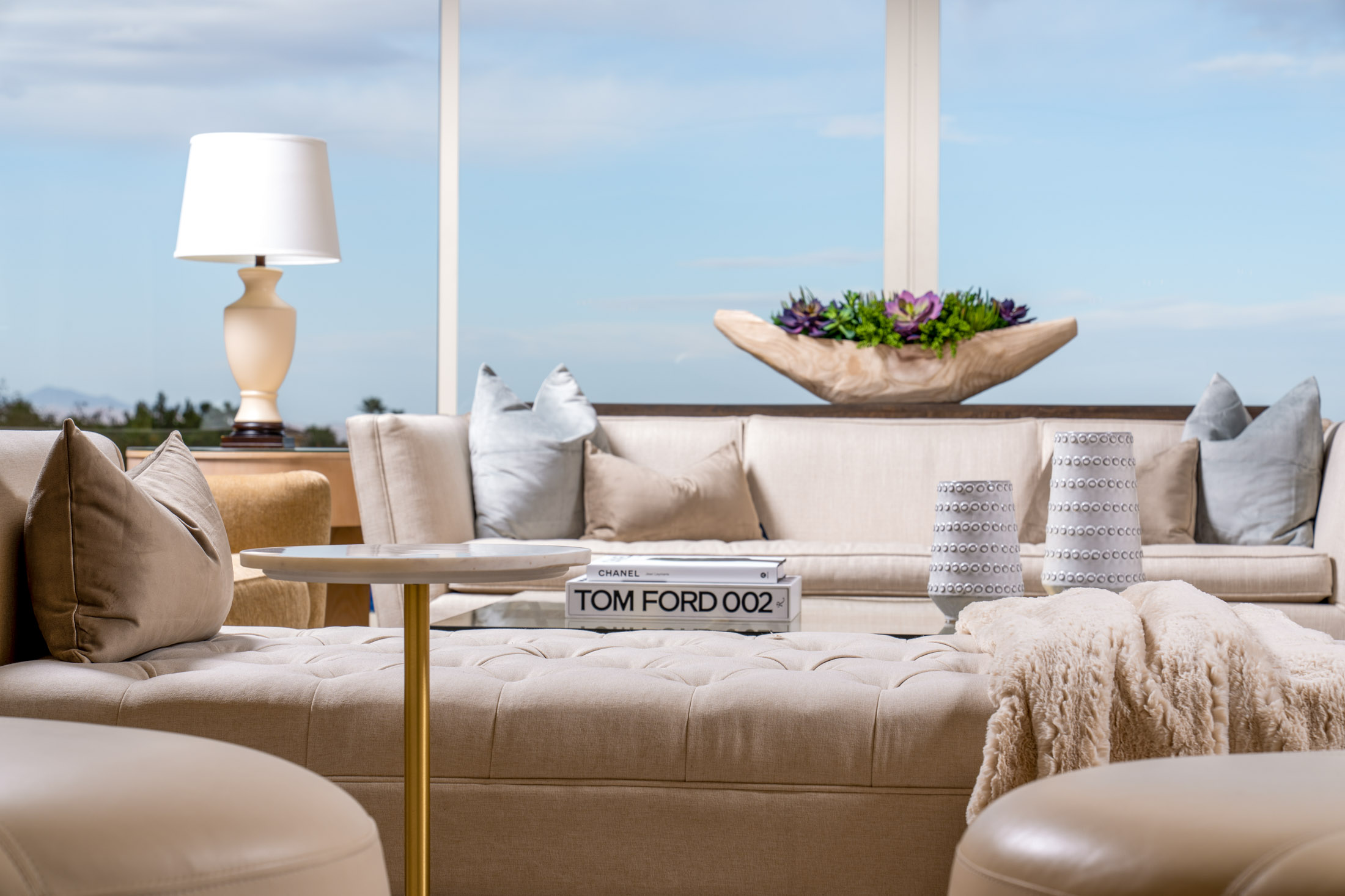 Luxury Home Staging A neutral seating area with a tufted ottoman in the foreground, styled with coffee table books and decorative vases. A succulent arrangement and a table lamp complete the cozy scene.