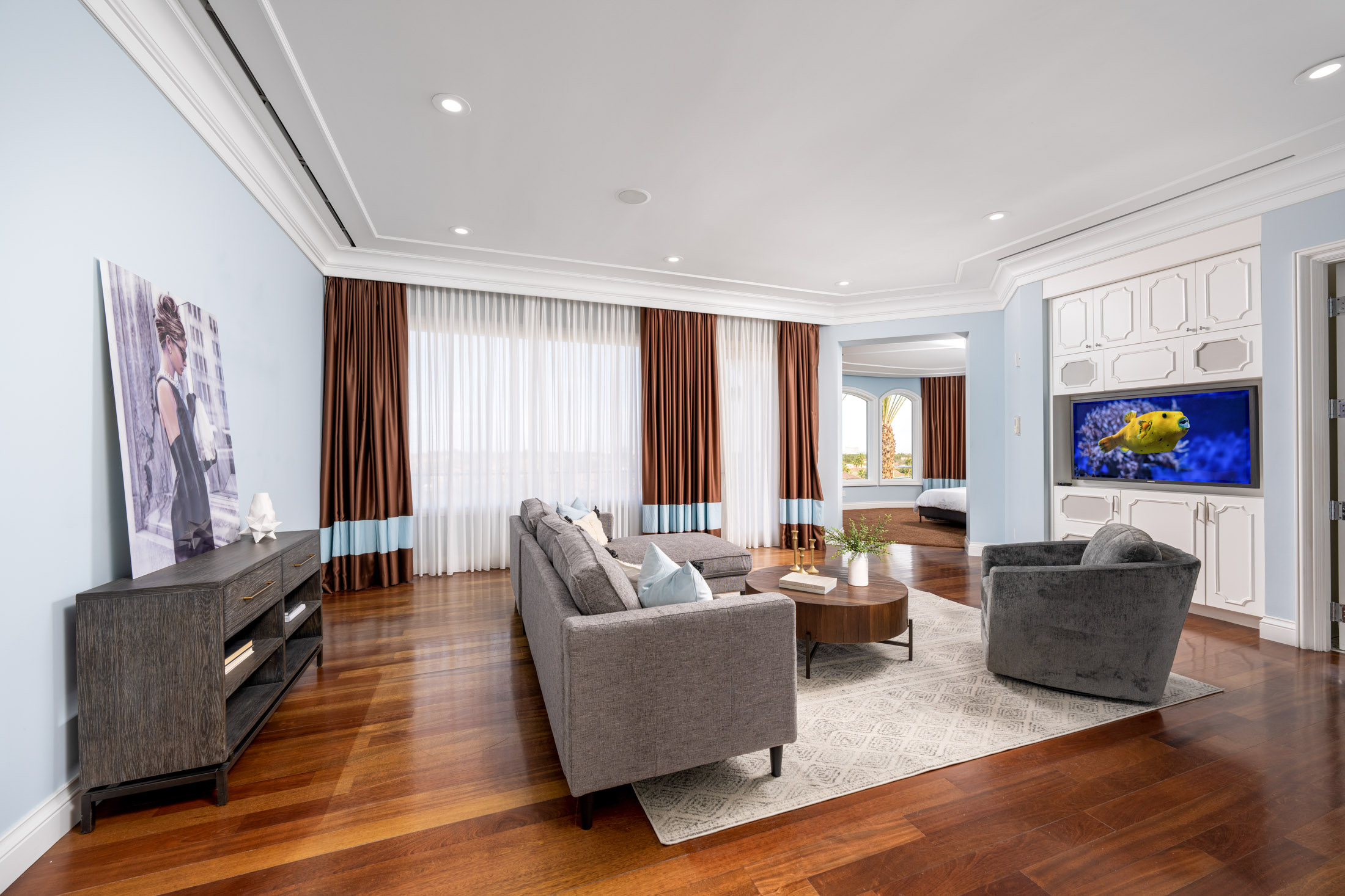 A wide living area with a gray sectional sofa, a round wooden coffee table, and rich brown curtains with a blue border. A large TV is built into cabinetry on the right, and an archway leads to a bedroom.