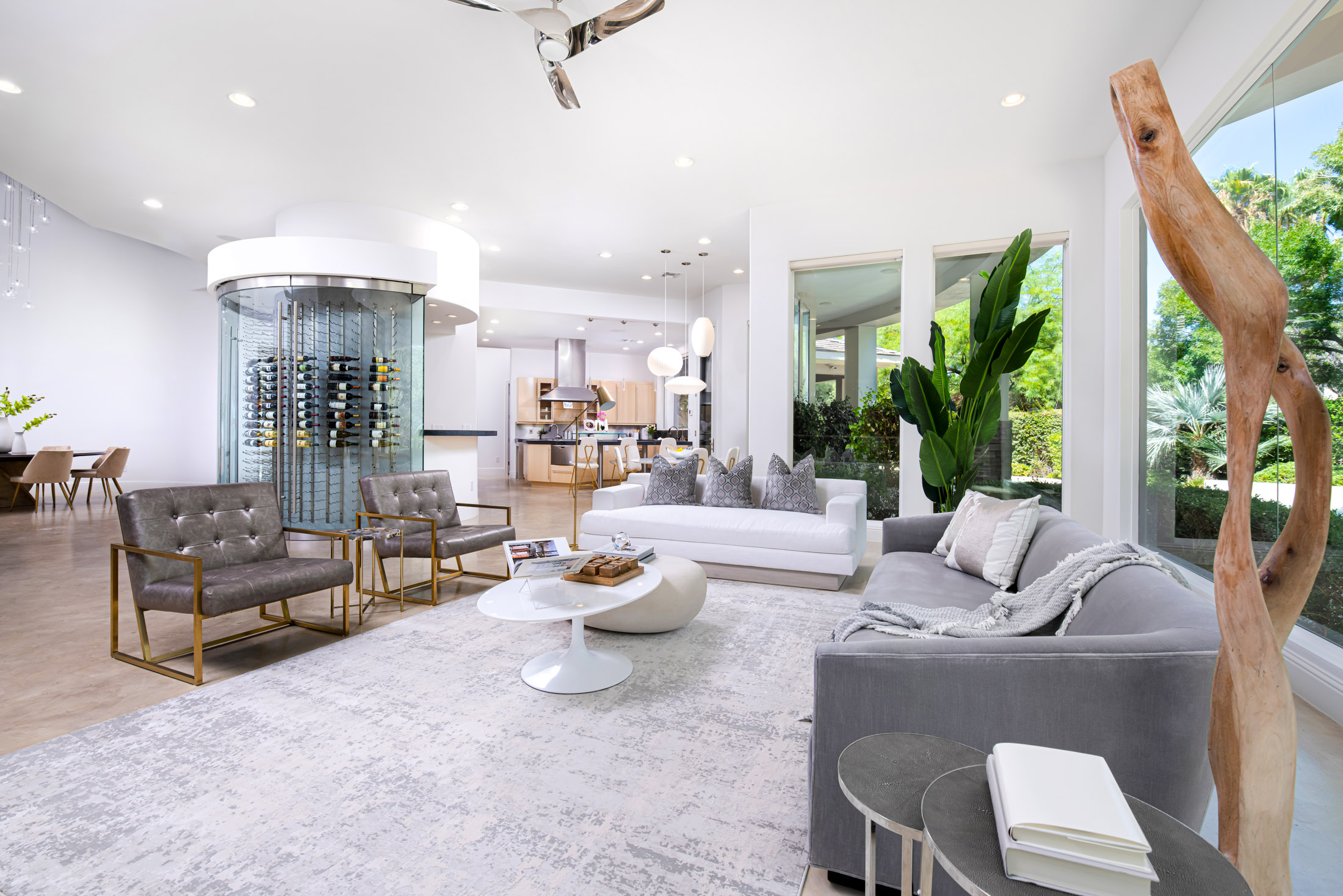 A fully staged living room with plush seating, gold-accented chairs, a statement wine display, and expansive windows bringing in natural light.