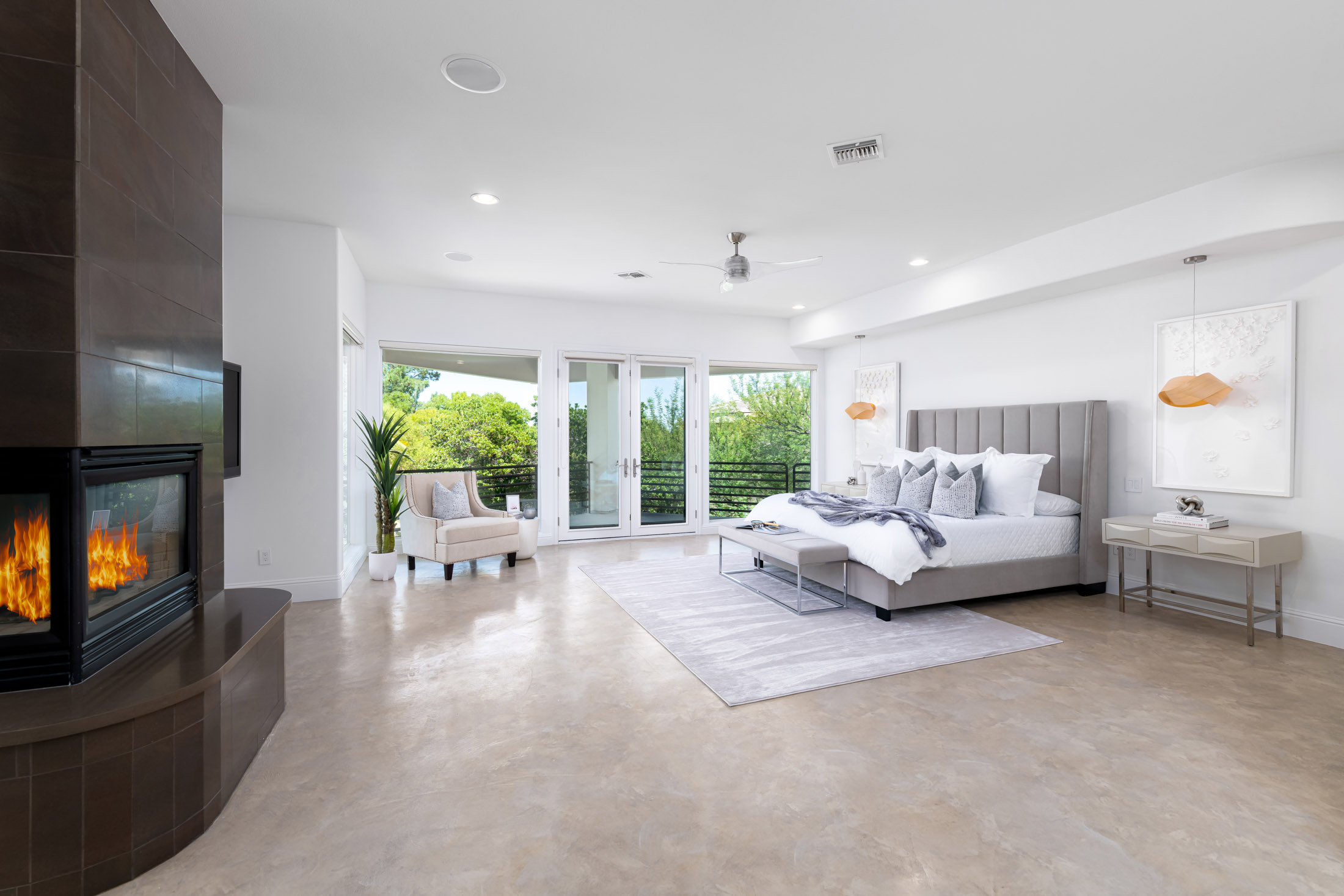 Las Vegas Home Staging A spacious master bedroom featuring a tufted gray bed, a cozy fireplace, a sitting area, and large glass doors leading to a balcony with lush greenery.