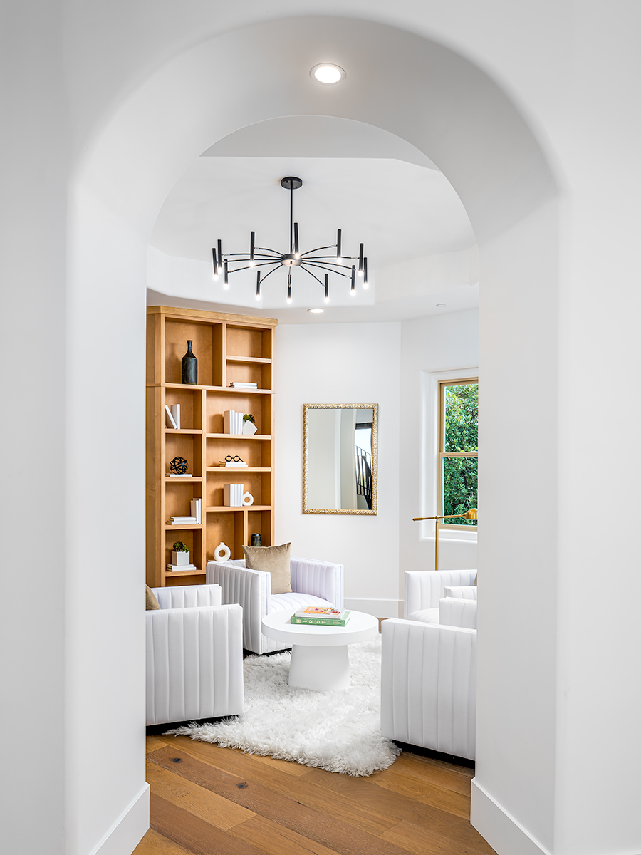 Cozy reading nook with white armchairs, a round white table, and a built-in wooden bookshelf filled with books and decorative items. There is a large window providing natural light, and a modern chandelier hangs from the ceiling, creating a comfortable and inviting space for relaxation.