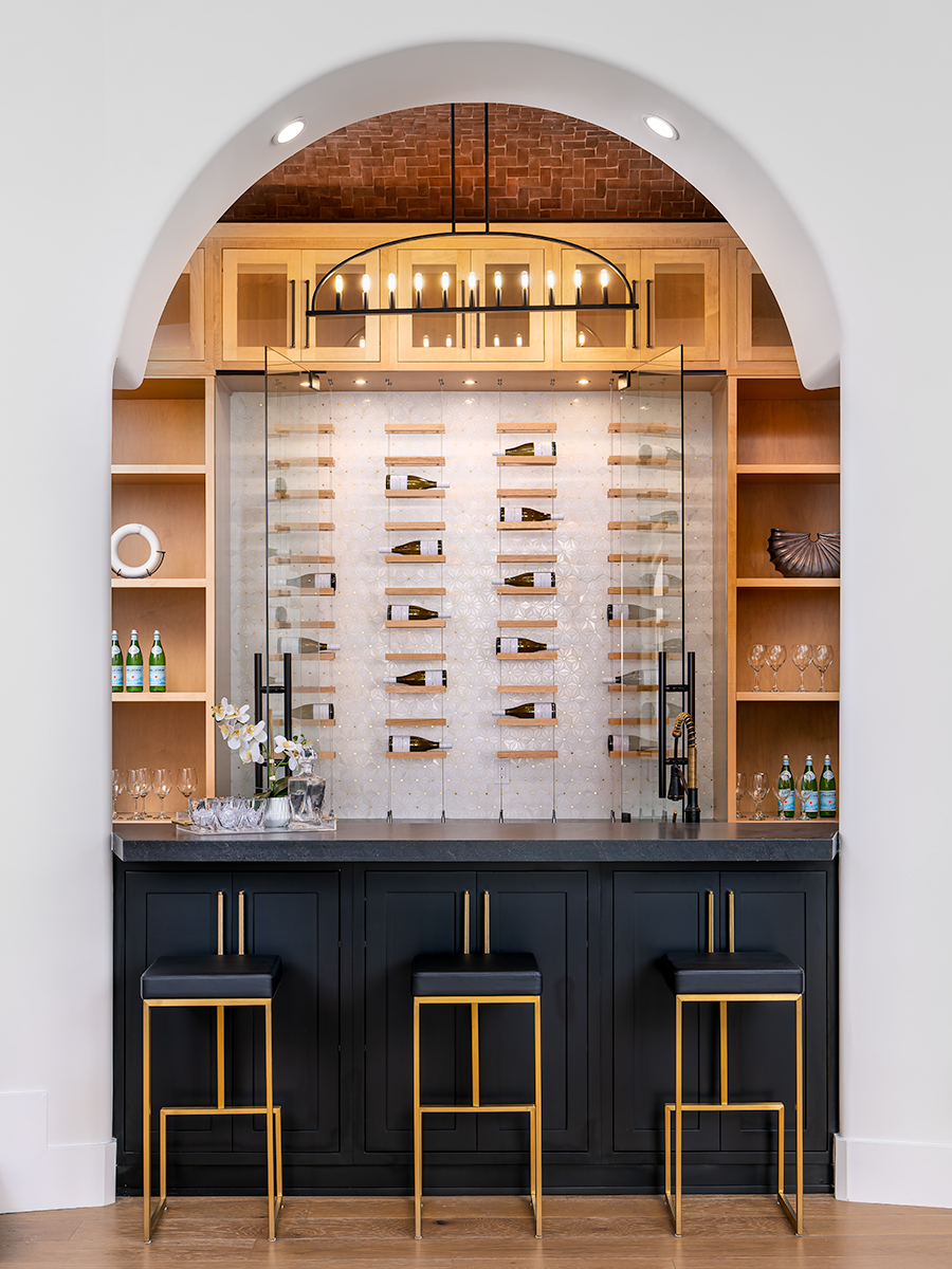 Stylish home bar with a black countertop, gold bar stools, and a wine display wall. Wooden shelves on either side are filled with glassware and decorative items, and a modern chandelier hangs above the bar, creating an elegant and sophisticated space for entertaining.