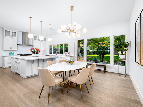A contemporary kitchen with a white island, elegant dining table, beige chairs with gold legs, and large windows showcasing lush greenery.