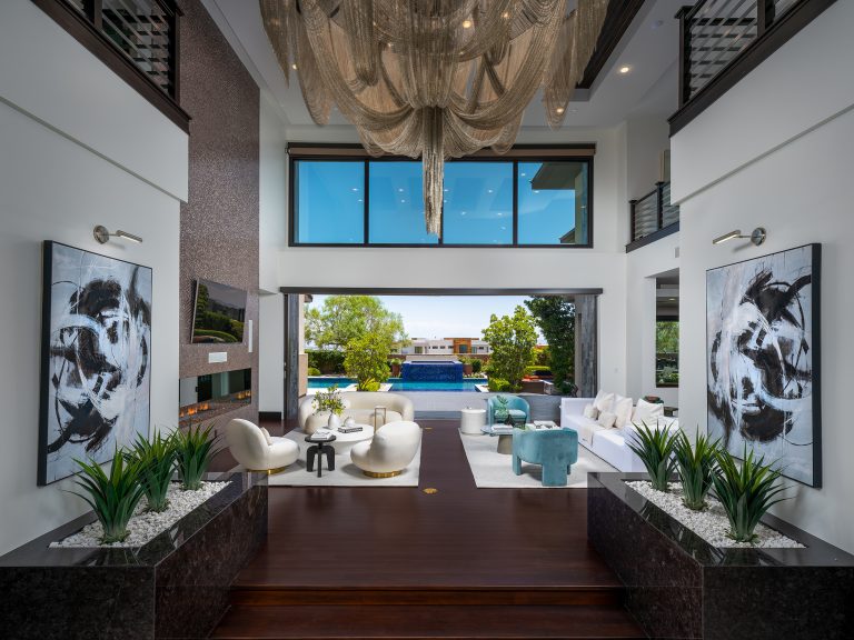 A modern living room with plush white and teal furniture, a sleek fireplace, and floor-to-ceiling sliding doors opening to a pool and lush outdoor space.
