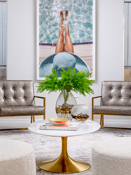 A stylish living area with tufted metallic chairs, a round white and gold coffee table, and a bold poolside-inspired artwork.