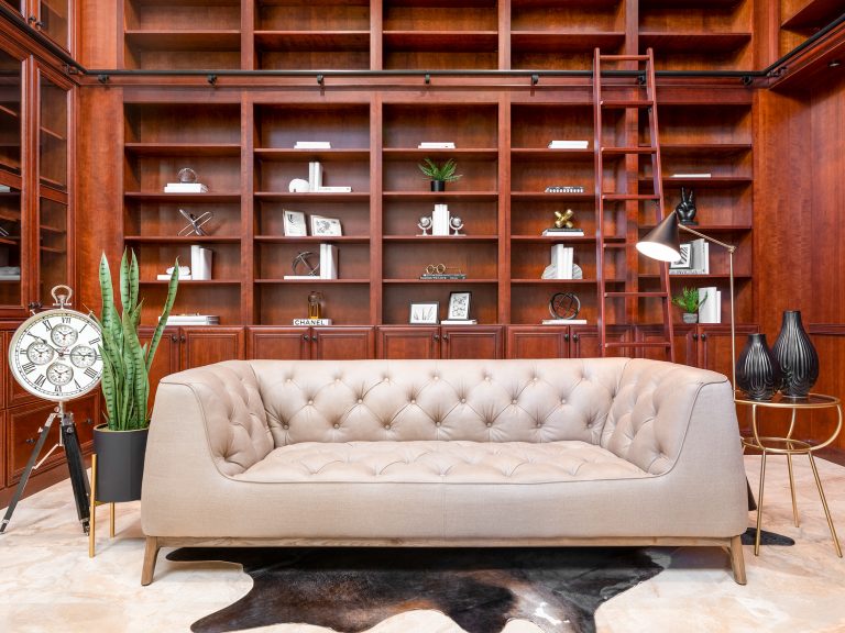 A tufted beige sofa in front of tall wooden bookcases, complete with a rolling ladder and various decorative items.