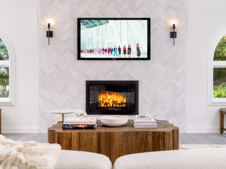 A living room centered around a herringbone-tiled fireplace with a rustic wood coffee table, and a TV mounted above the fire.