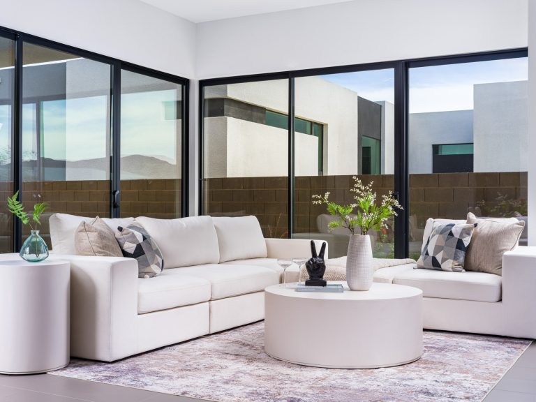 A white sectional sofa with geometric pillows by floor-to-ceiling windows, centered around a round coffee table.