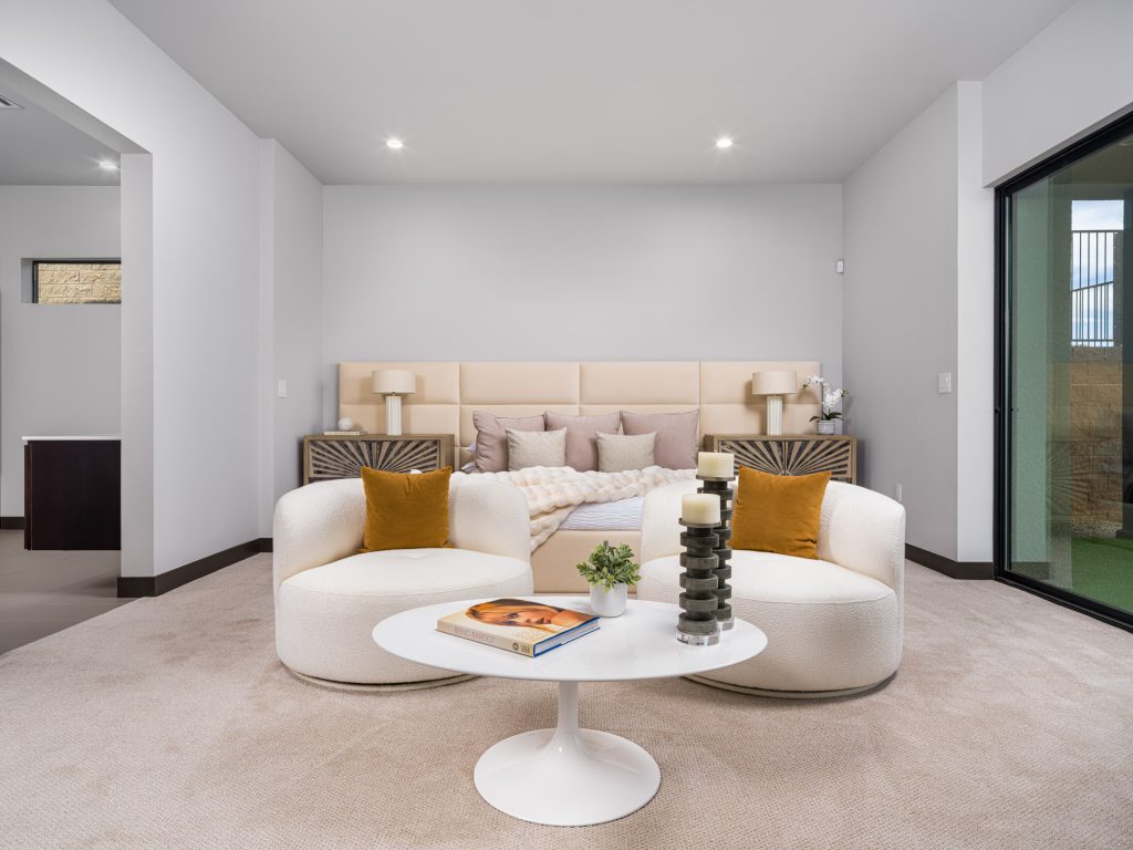 A neutral-toned bedroom with a padded headboard, two curved chairs, and a white coffee table near sliding glass doors.