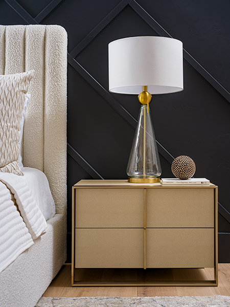 A stylish bedroom nightstand with a textured bed frame, a glass and gold lamp, and a dark accent wall with geometric detailing.