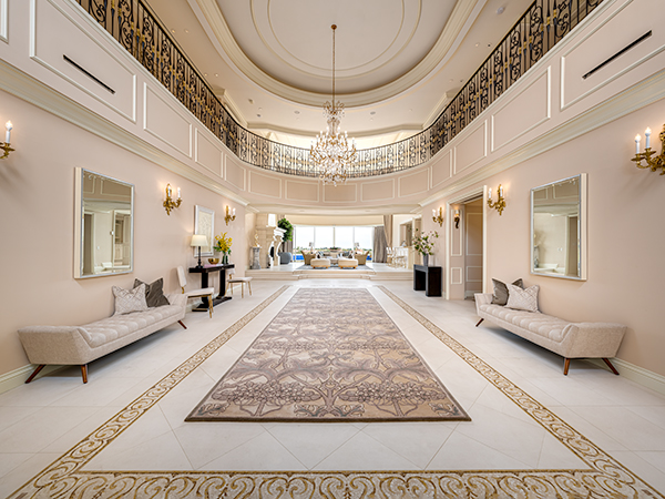 A grand foyer with a high ceiling, intricate iron railings, a crystal chandelier, and a luxurious rug leading to an open living space.