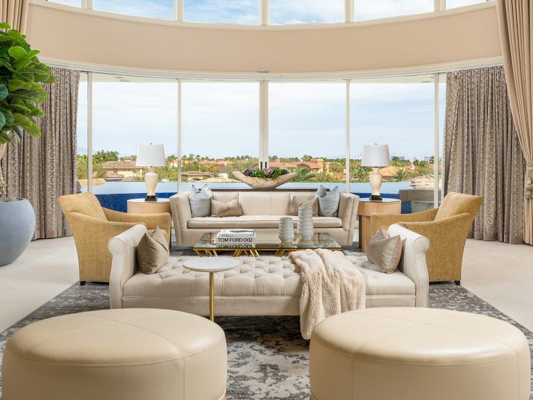 A bright living room with large curved windows, neutral-toned seating, and a pool visible through the glass.