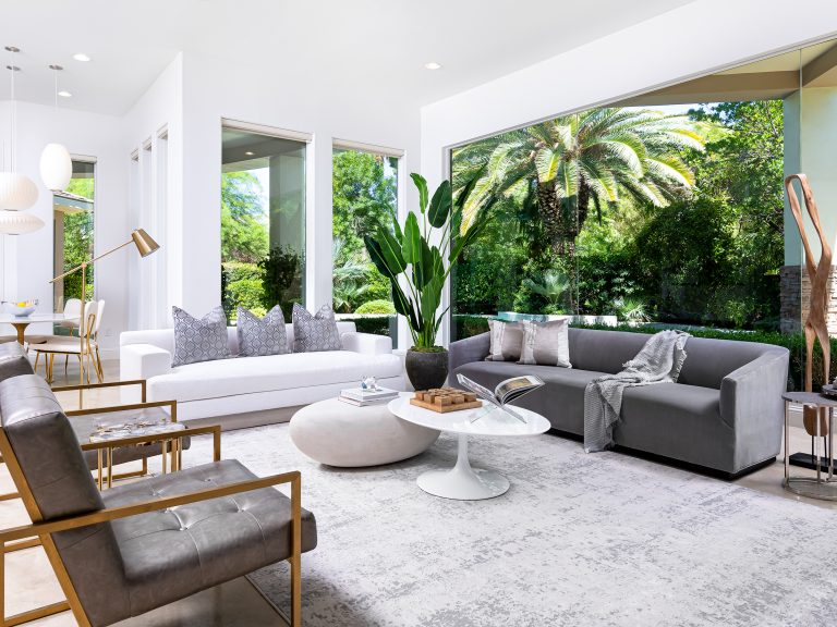 A bright living area with large windows, white and gray sofas, and gold-accented chairs, showcasing lush palm trees outside.